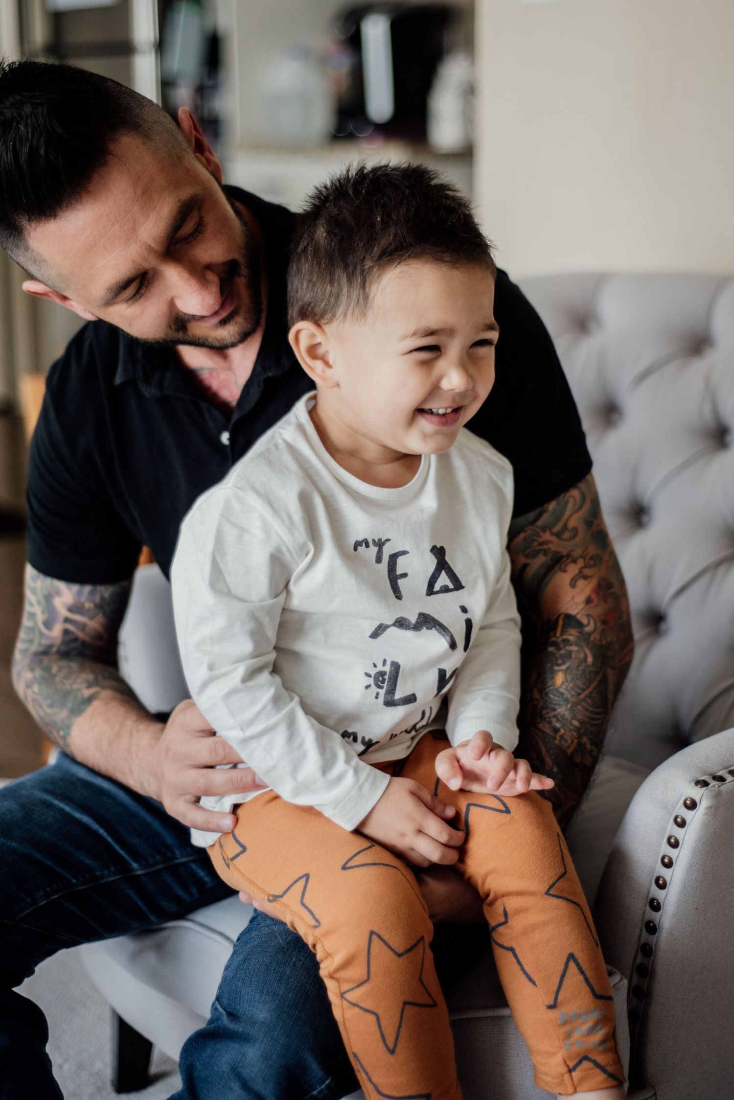 A parent and child share a joyful moment together on a gray couch, with the child wearing patterned orange pants.