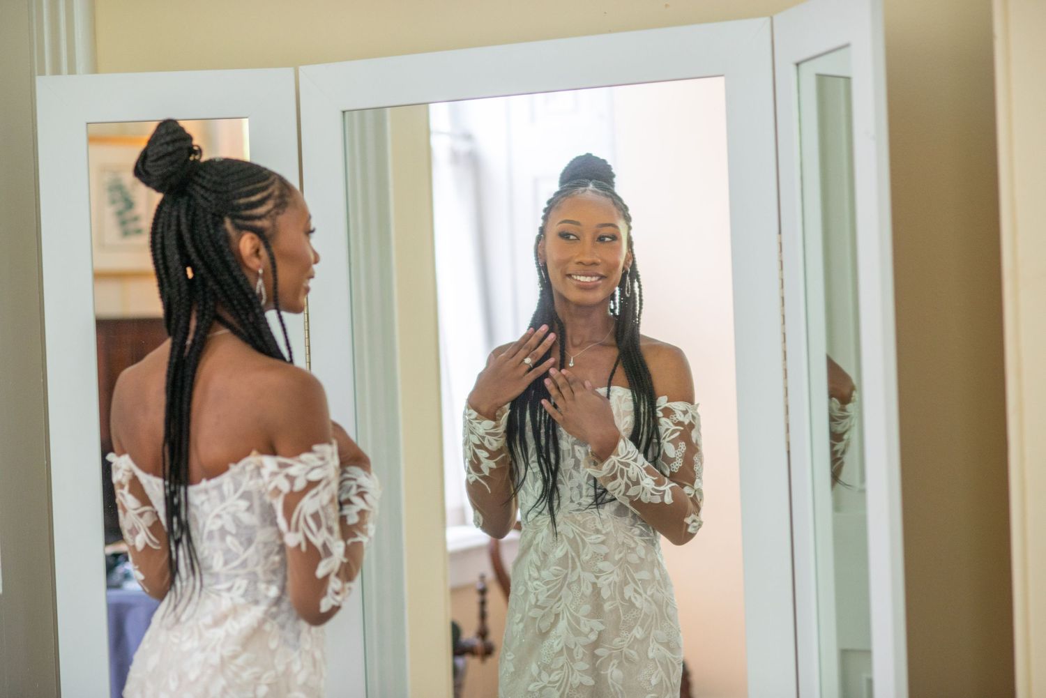 A bride in a detailed white wedding dress with off-shoulder lace design stands looking at her reflection in a mirror.