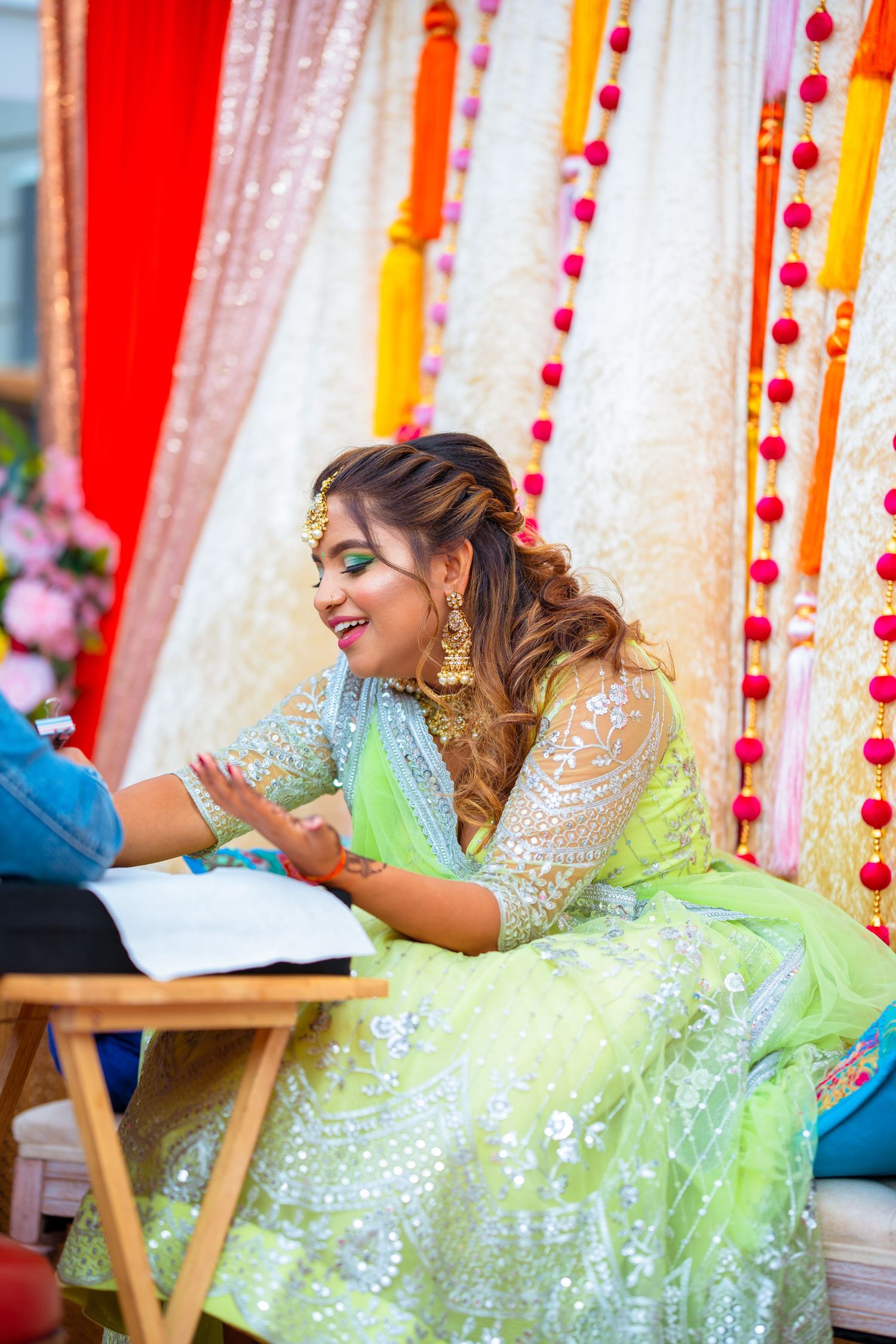 Person wearing lime green embellished lehenga sits during traditional Indian mehndi ceremony with colorful backdrop.
