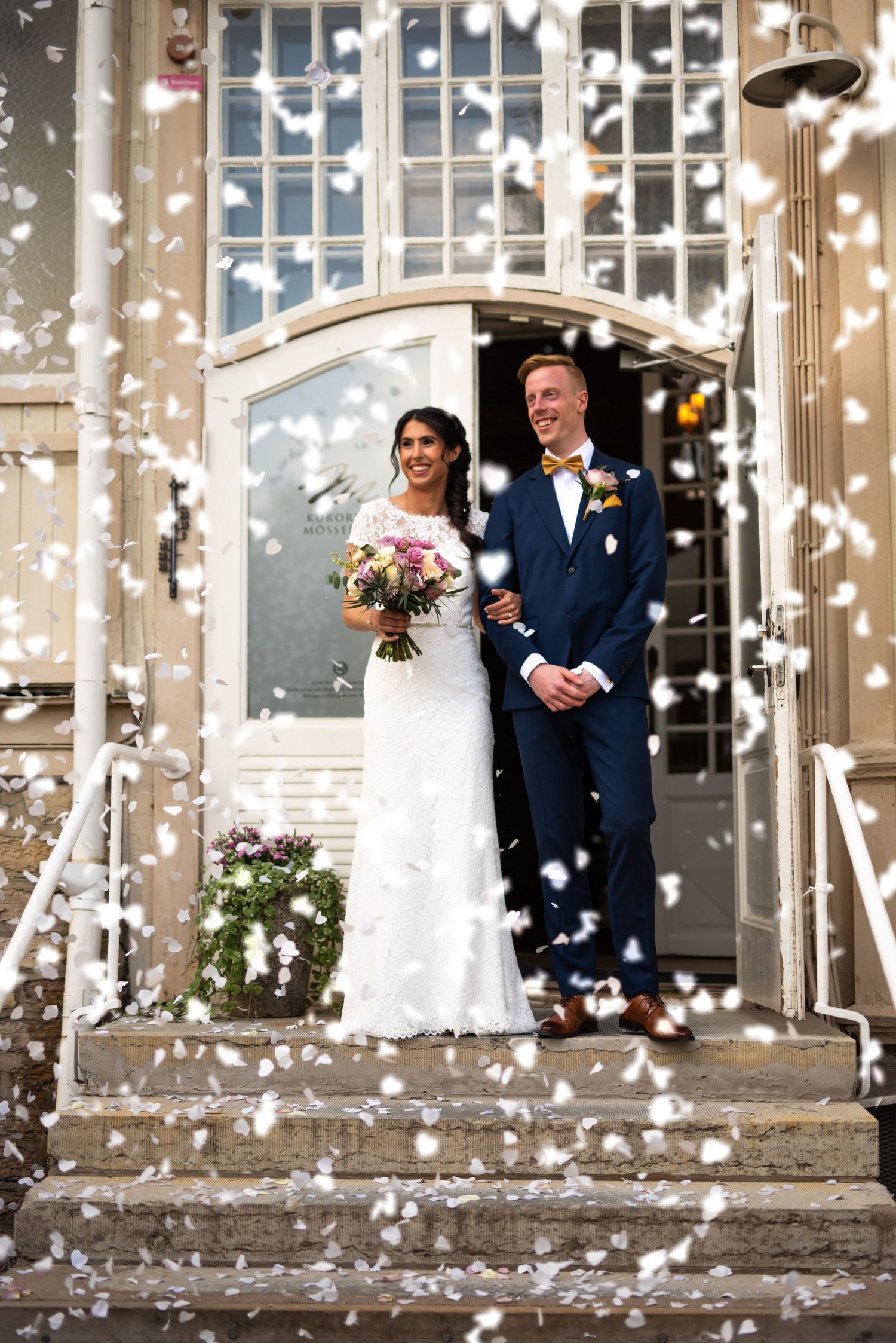 Newlywed couple standing on steps in a shower of white confetti outside a building.