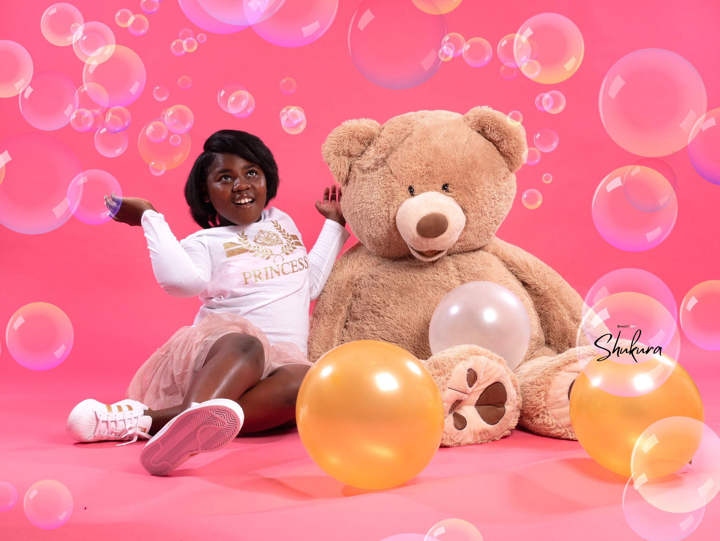 Child playing with large teddy bear and balloons against pink background with floating bubbles.