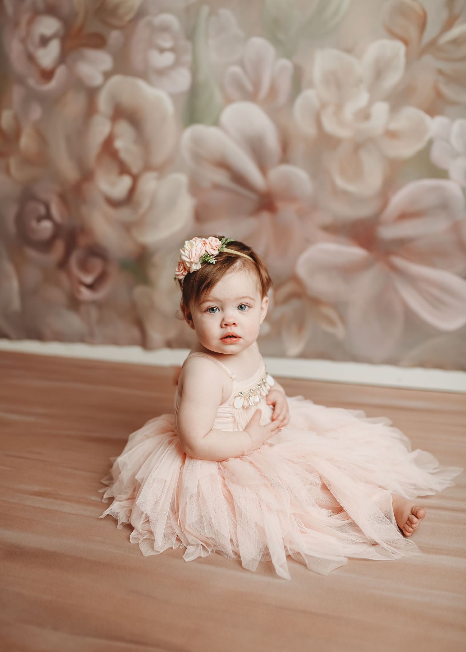 Baby in pink tulle dress lying on wooden floor with rose patterned backdrop.