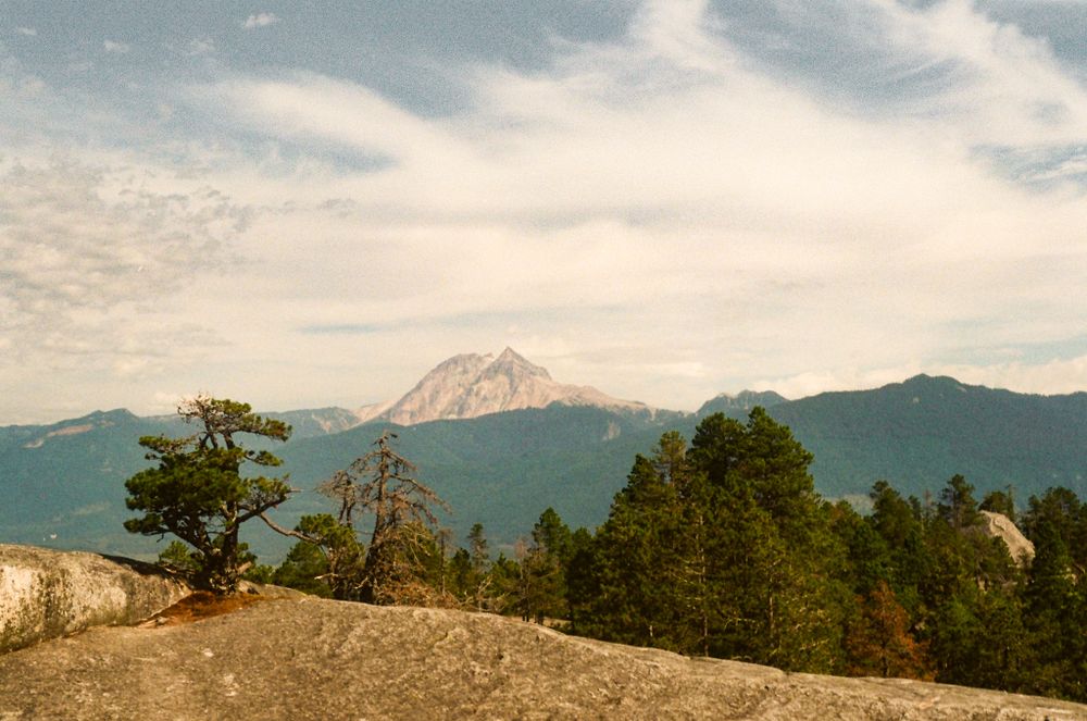 Dramatic mountain peak rises above rocky terrain with sparse pine trees against cloudy sky.