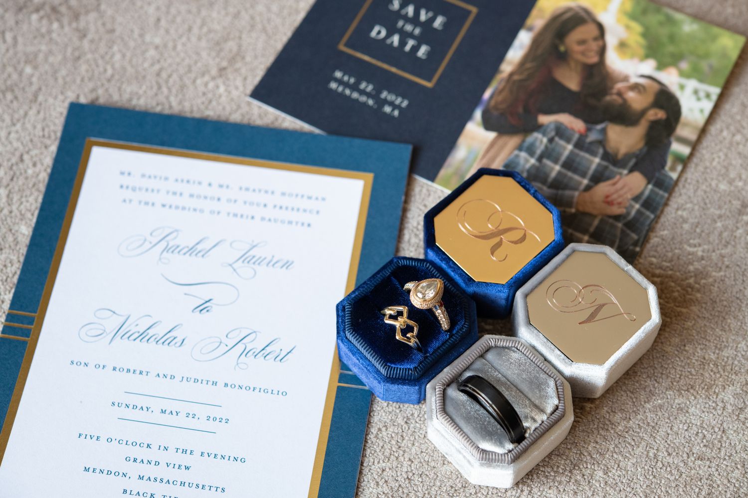 Wedding invitation suite with blue and gold details alongside ring boxes on grey fabric.