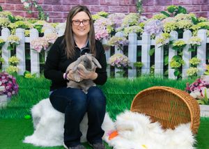 Person holds a bunny while sitting with easter decorations and white picket fence backdrop.