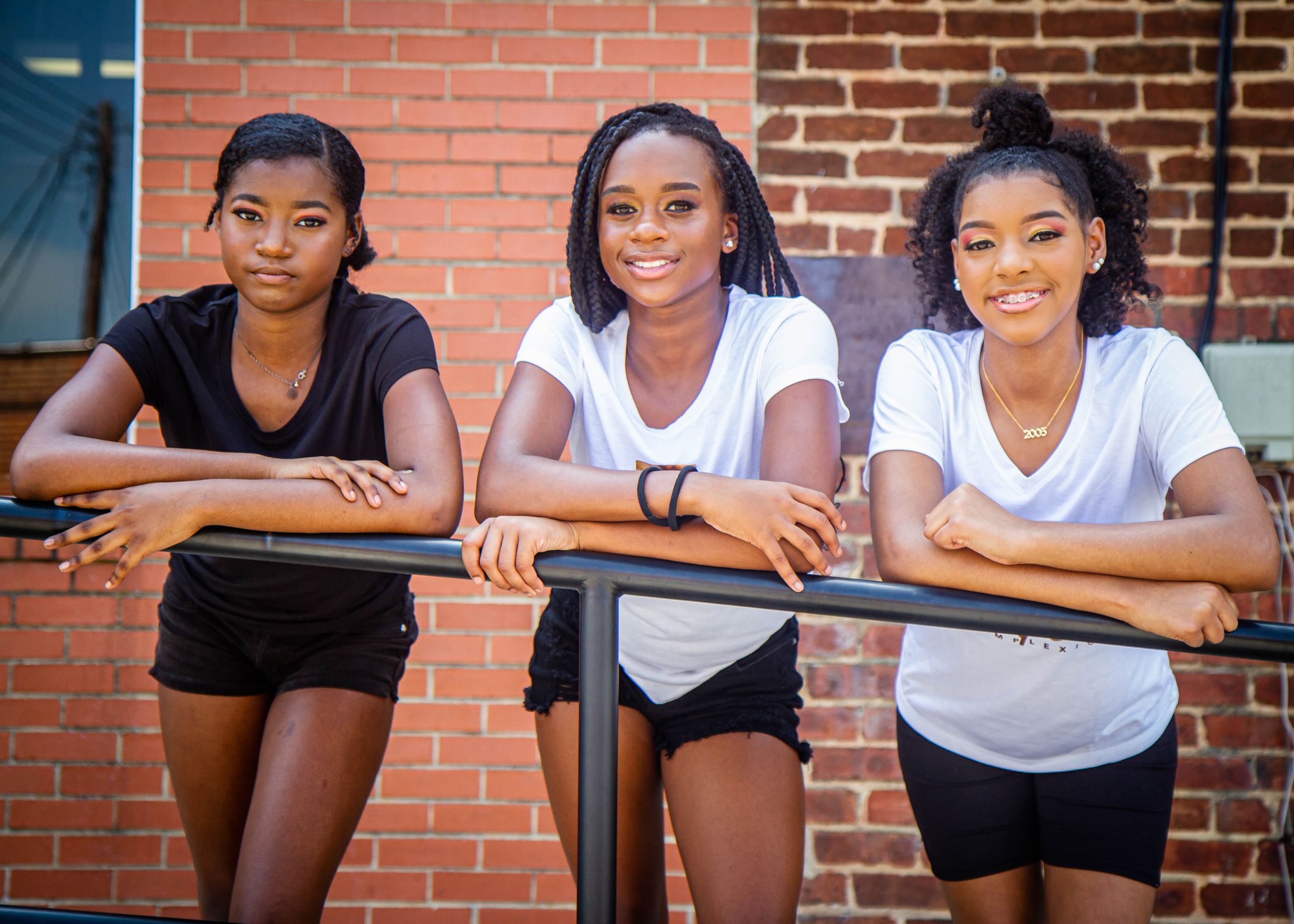 Three athletes in workout attire leaning on railing against brick wall background.
