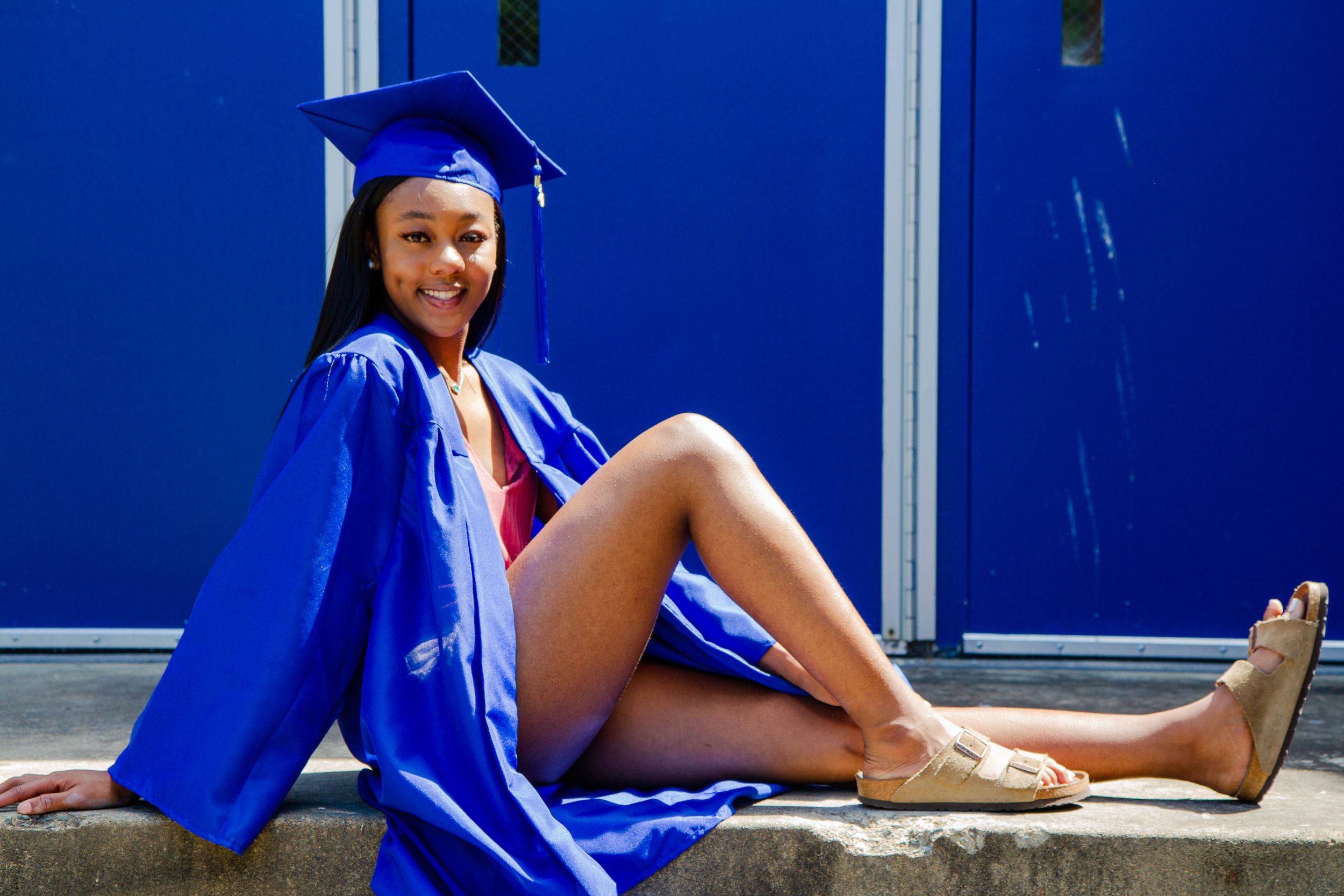 A graduate in blue cap and gown poses sitting against a bright blue wall.