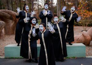 Performers in black robes and white face paint pose dramatically in an outdoor setting.