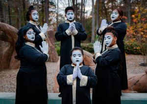 Mime performers in black robes and white face paint gesture dramatically in a theatrical pose.