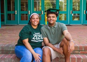 Two graduates in green caps and gowns sit on brick steps in front of glass doors.