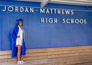 A graduate in blue cap and gown poses in front of Jordan-Matthews High School sign.