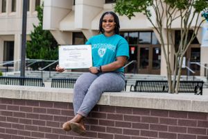 A joyful graduate displays their diploma while sitting on a brick wall outside a courthouse.