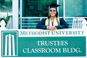 Graduate poses in front of Methodist University Trustees Classroom Building sign wearing cap and gown with honors cords.