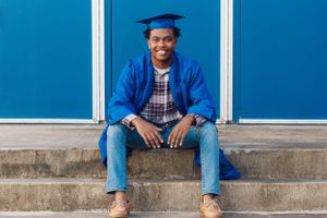 Graduate in blue cap and gown sits on concrete steps against blue wall.