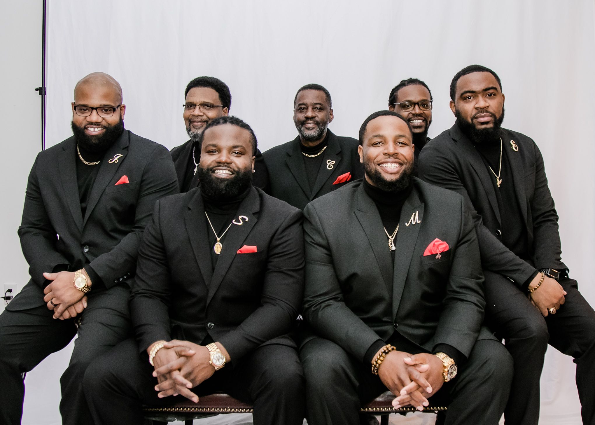 Group portrait of men in matching black suits with red pocket squares sitting in formal arrangement.