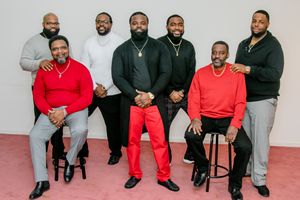 Group photo of men wearing coordinated outfits in black and red seated against light background.