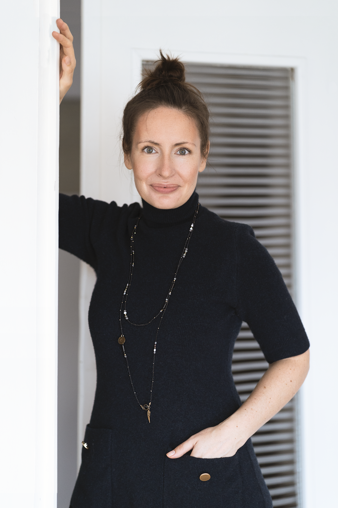Portrait against white wall wearing black turtleneck and long necklace.