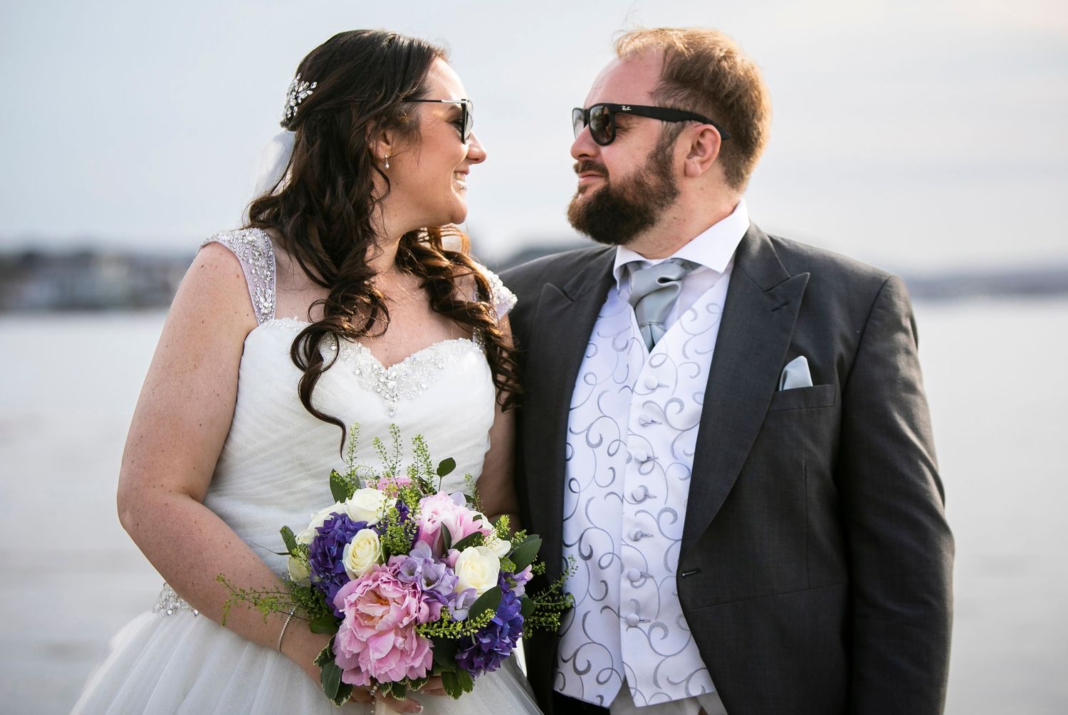 Romantic waterfront wedding portrait with floral bouquet and formal attire.