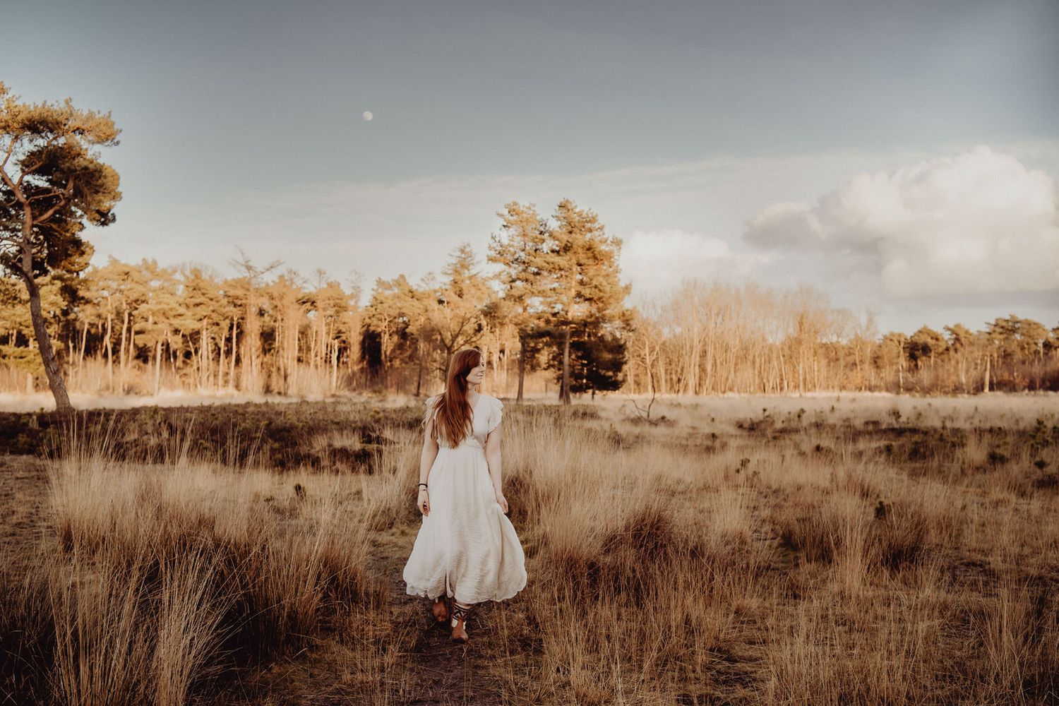 A white dress stands out against golden grass and autumn trees at sunset.