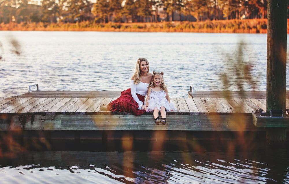 Two people in dresses sit together on a wooden dock by a lake at sunset.
