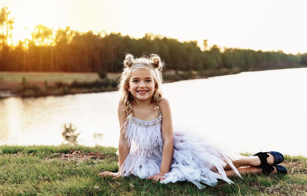 Small child in a white dress sitting on grass by a lake during golden hour.
