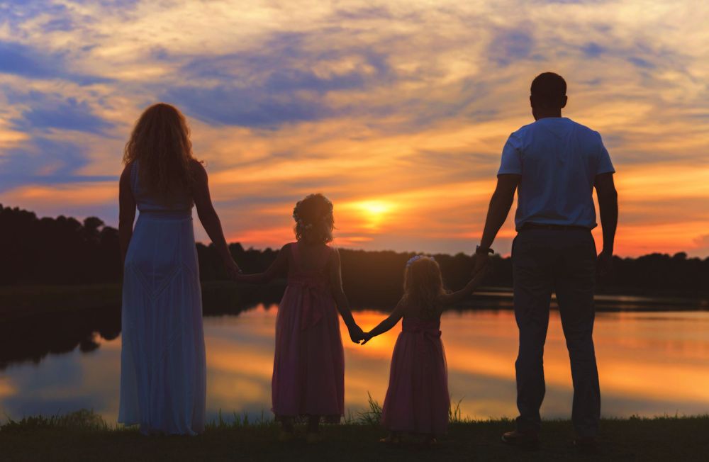 Silhouettes of a family against a dramatic orange and blue sunset sky by a lake.