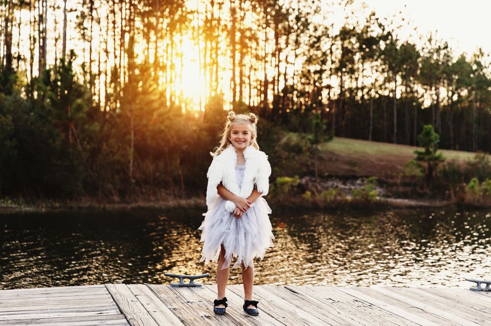 Young child in white dress standing on dock at sunset.