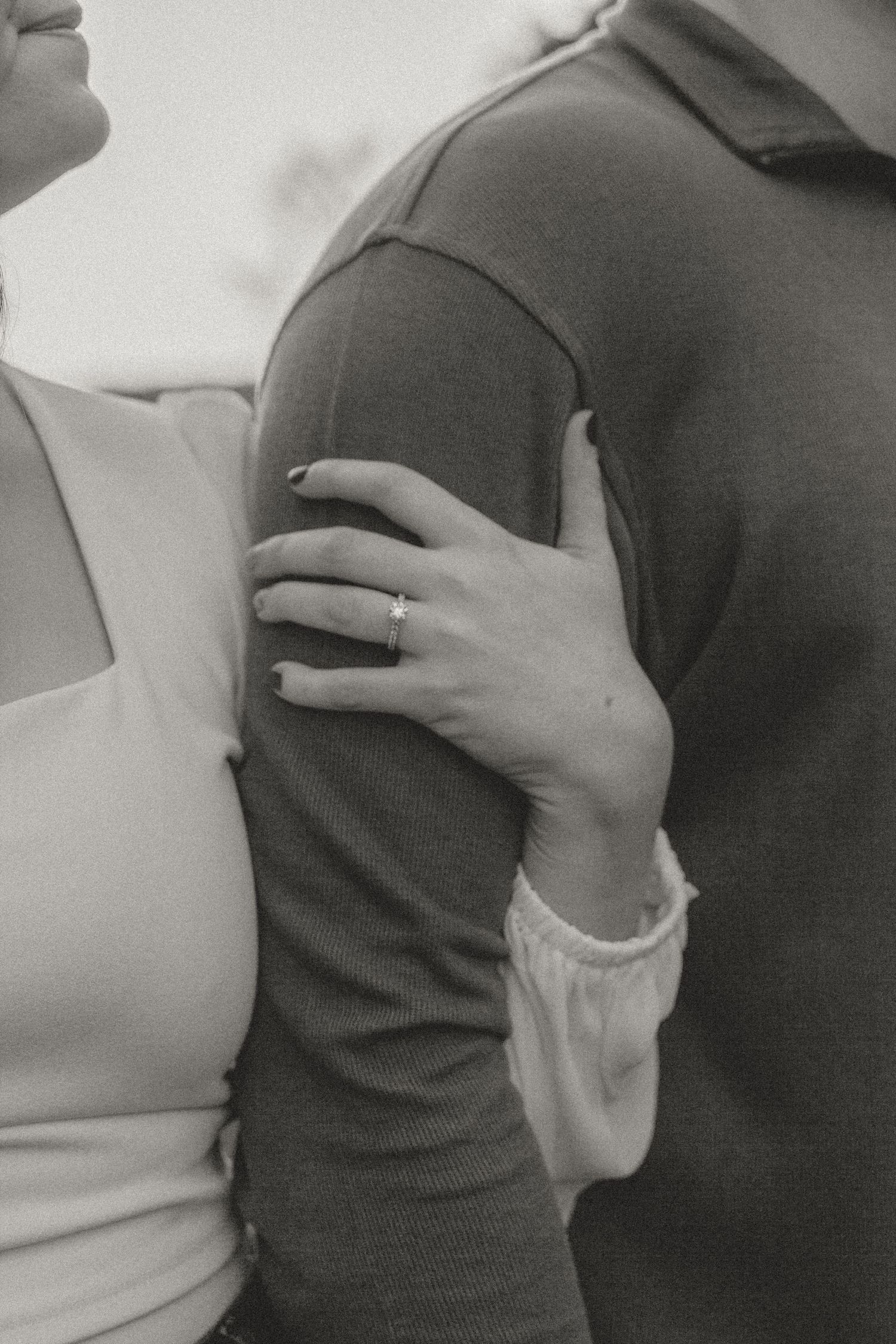 A close-up black and white photo of hands embracing with a wedding ring visible.