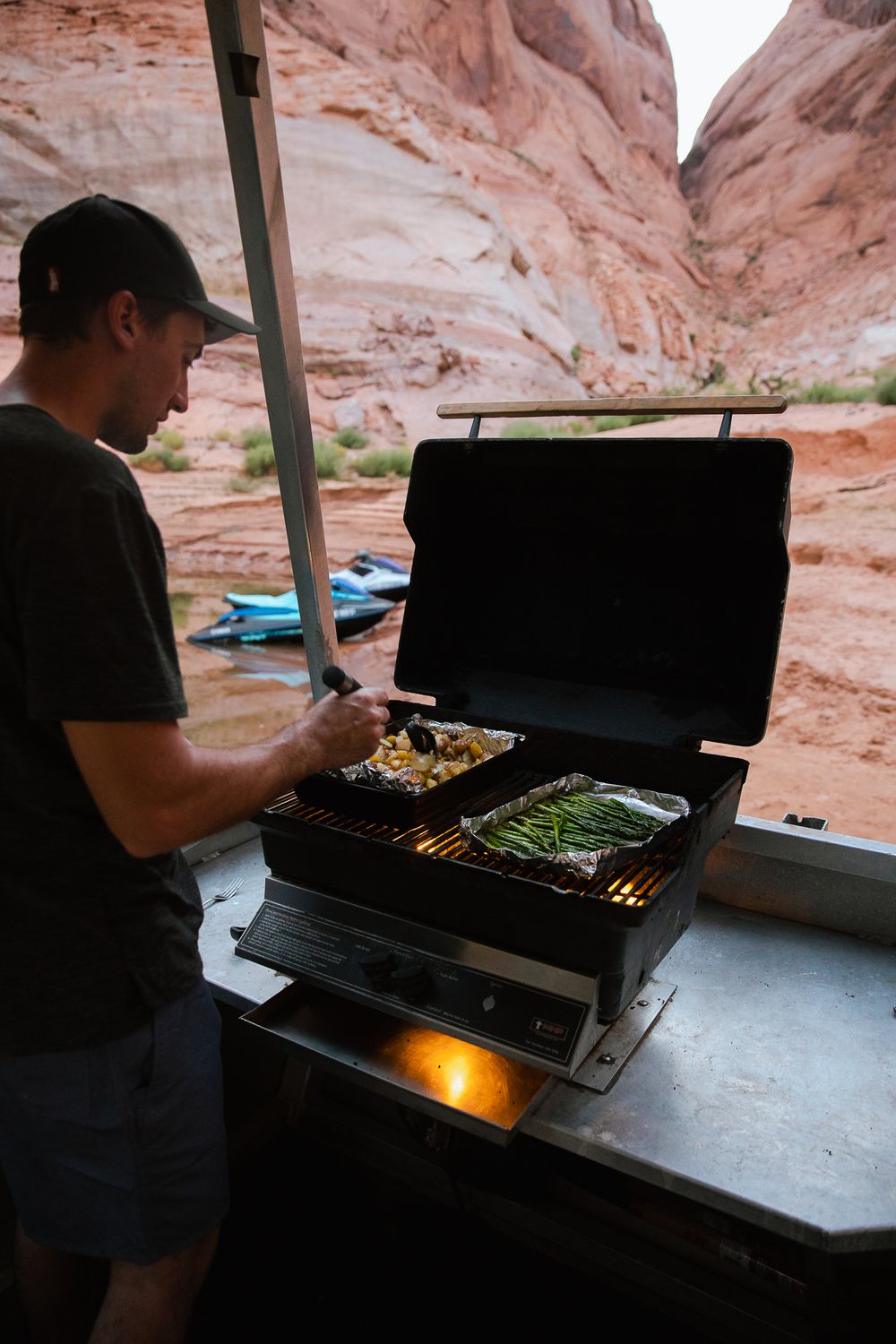 man grilling food over small grill on back of houseboat beached on shore with red rocks and jetski surrounding