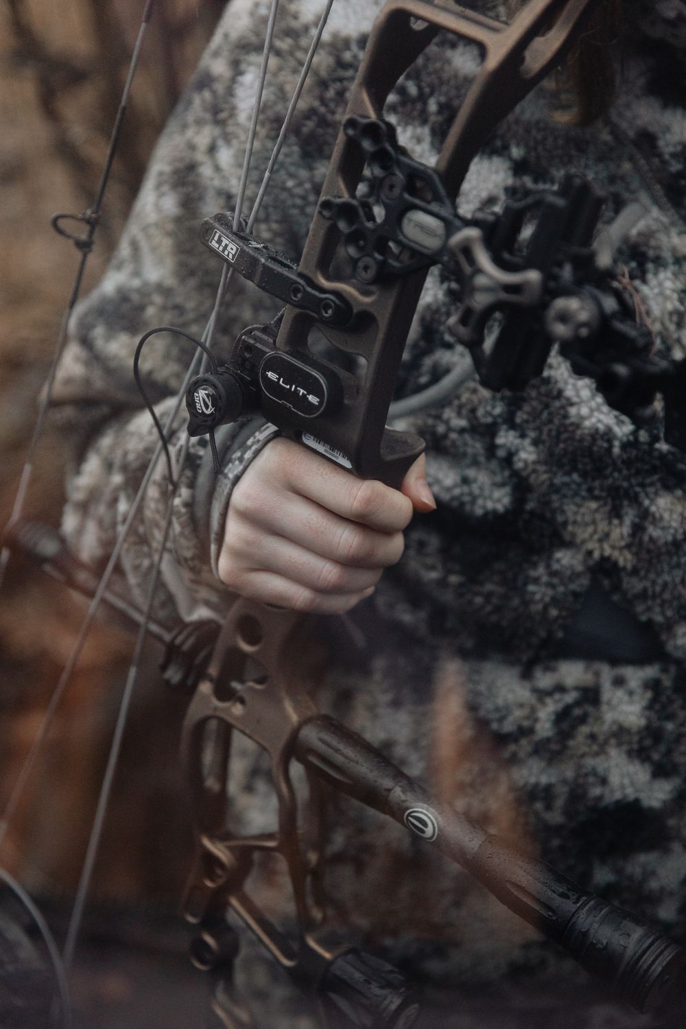 close up shot of person holding bronze bow in hand wearing all camo with red and brown colors around