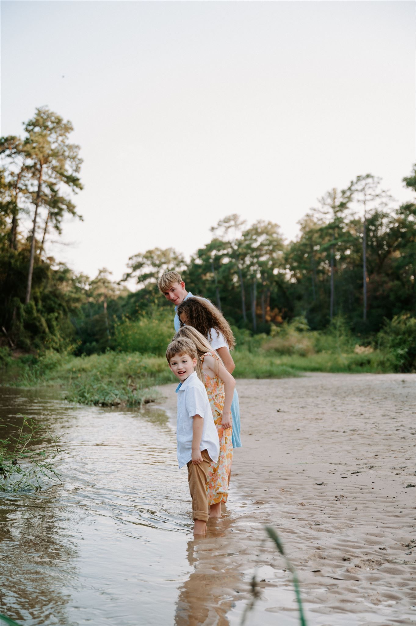 People stand in shallow water at the edge of a peaceful river surrounded by trees.