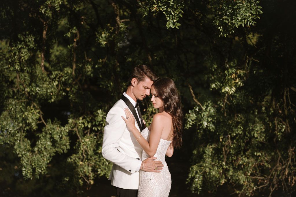 Newlyweds share an intimate embrace surrounded by lush green foliage in an outdoor setting.