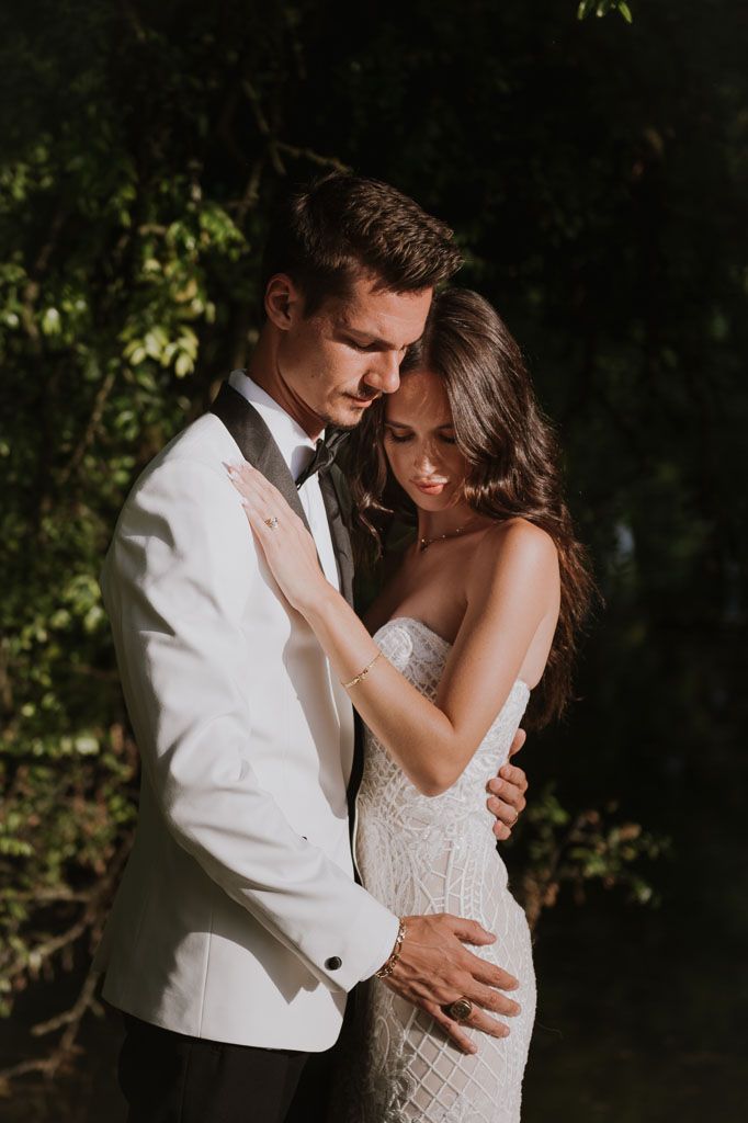 Romantic wedding photo capturing intimate embrace with bride in fitted lace gown and groom in white tuxedo.