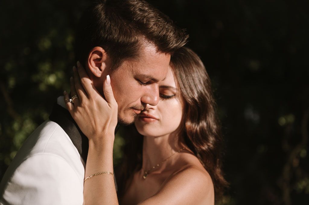 Tender moment between newlyweds captured in warm sunlight with natural greenery background.