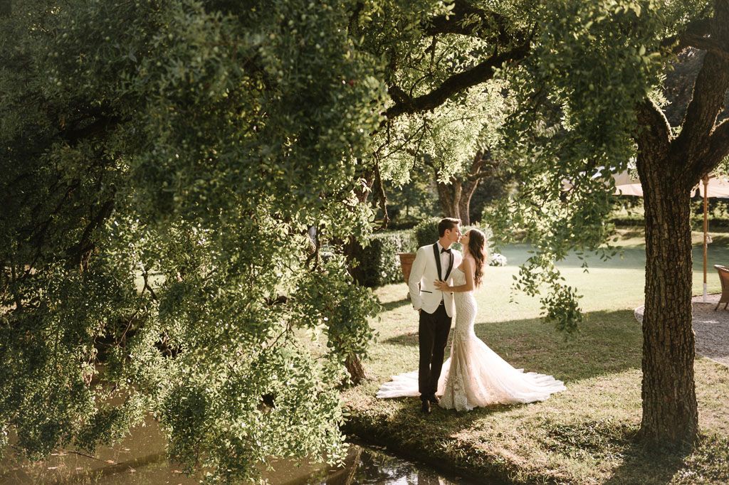 Dreamy wedding photo of couple standing beneath sunlit trees in a garden setting.