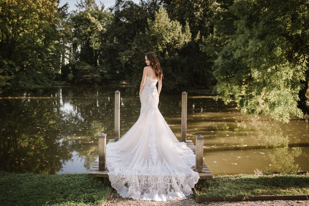 Beautiful bride in lace mermaid gown standing on wooden dock over tranquil water.