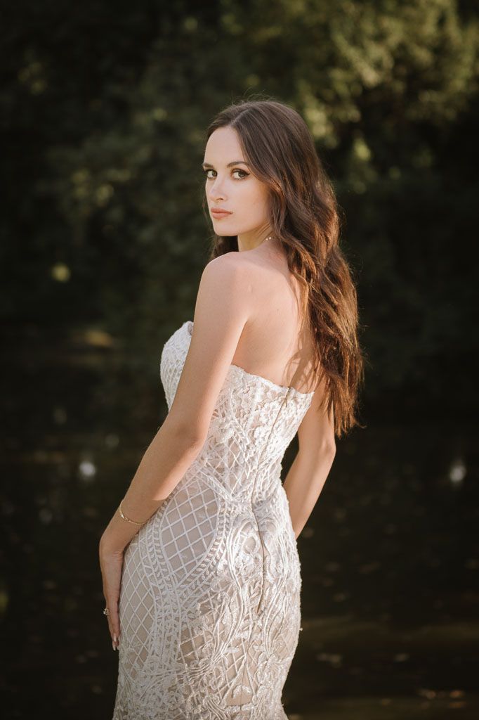 Elegant bride wearing strapless lace gown poses against natural backdrop during sunset photoshoot.