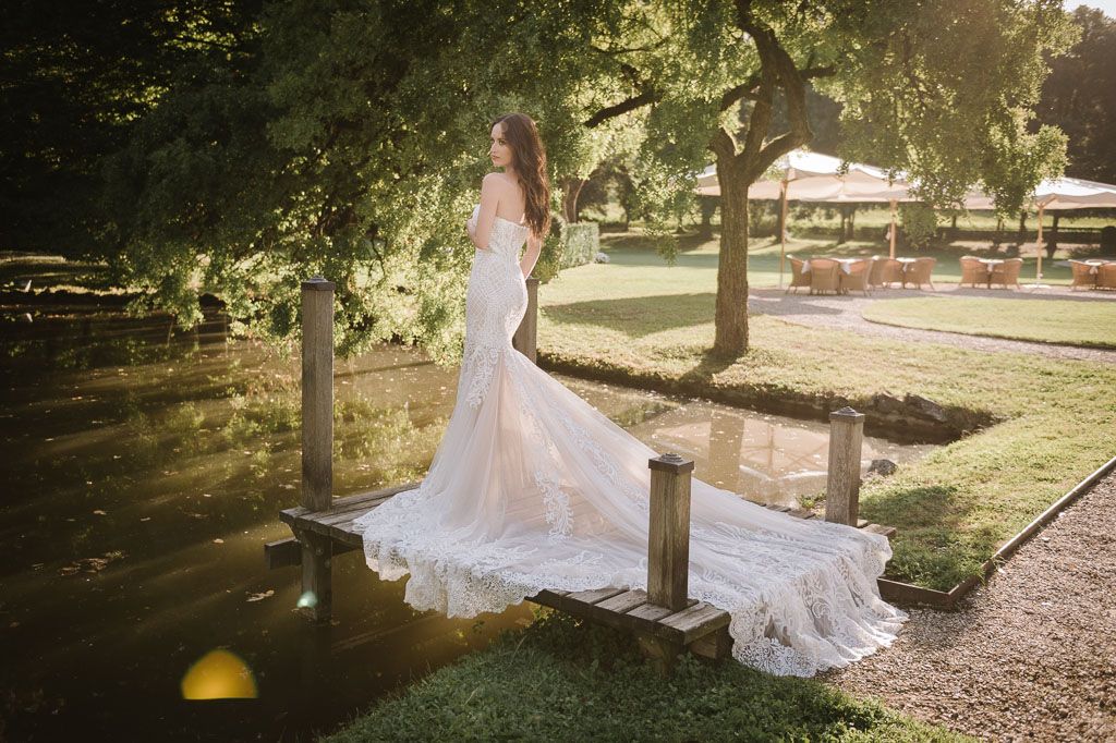 Bride in fitted mermaid wedding dress with long train poses on garden path during golden hour.