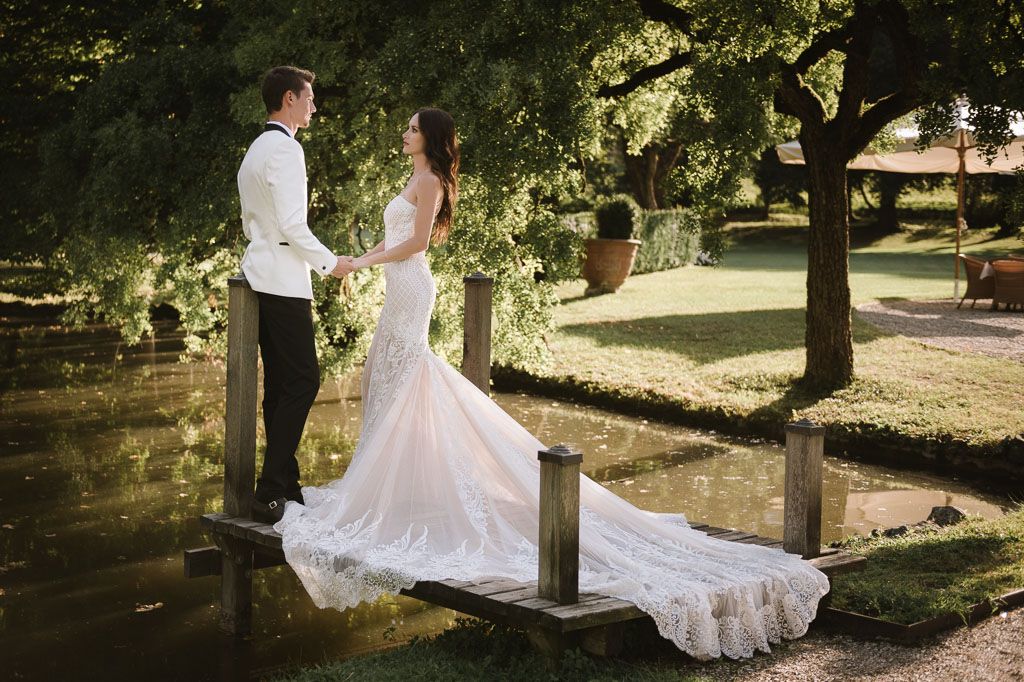 Newlywed couple in white formal attire sharing romantic moment on garden bridge.
