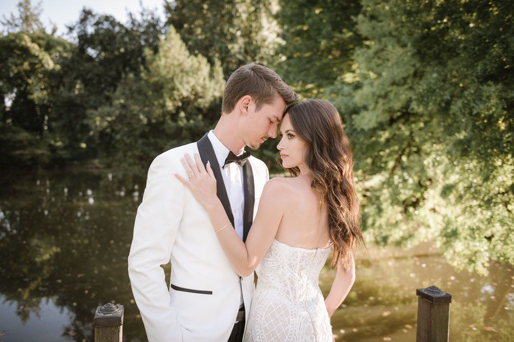 Intimate portrait of couple in white wedding attire embracing by pond in garden setting.