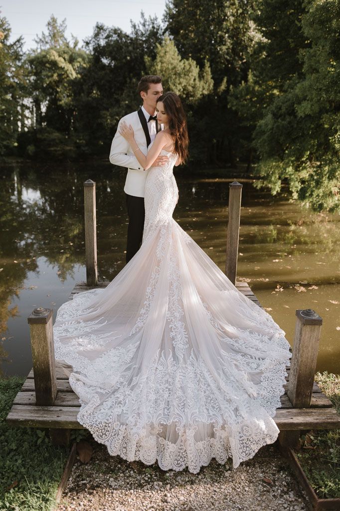 Romantic wedding couple embraces on wooden dock as bride's elegant lace mermaid gown trails behind them.