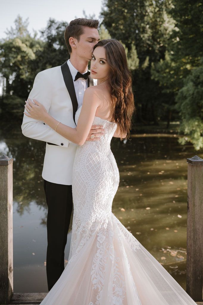 Stylish bride and groom share intimate moment on dock wearing white tuxedo and fitted lace wedding dress.