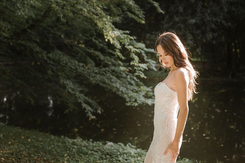 Sun-kissed bride in fitted lace dress poses against lush green forest backdrop.