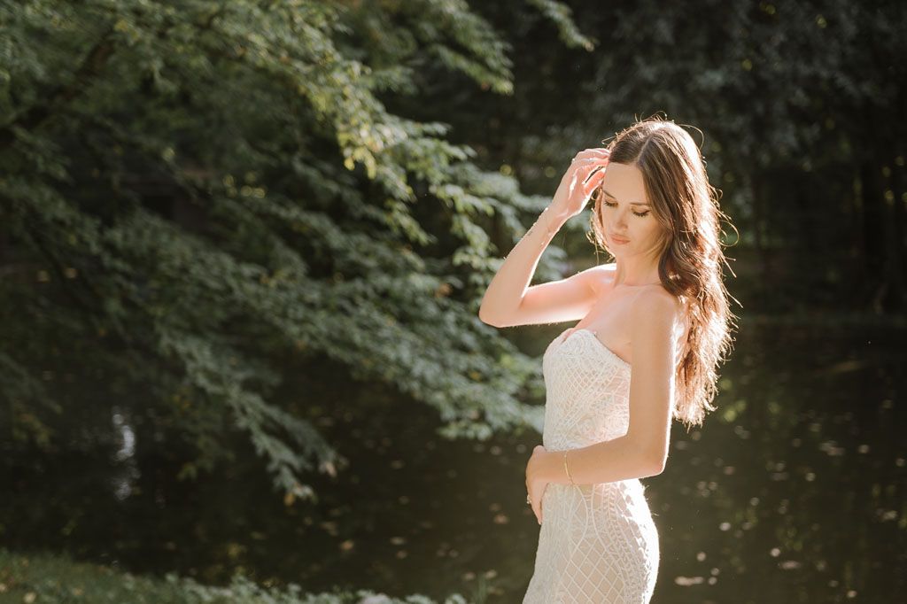 Elegant bride in two-piece wedding dress stands thoughtfully against forest scenery.