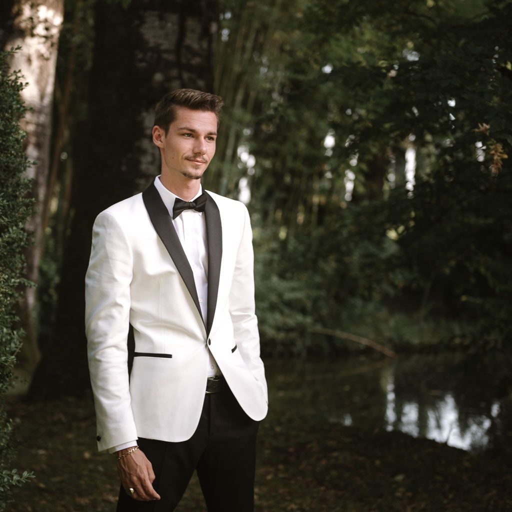 Groom in crisp white tuxedo jacket and black bow tie poses by tranquil pond.