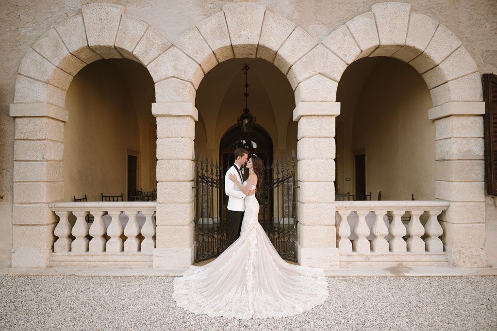 A romantic wedding photo captured under elegant stone archways with a flowing white dress train.