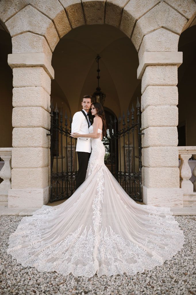 Wedding couple poses in formal attire under stone archway with dramatic train detail.