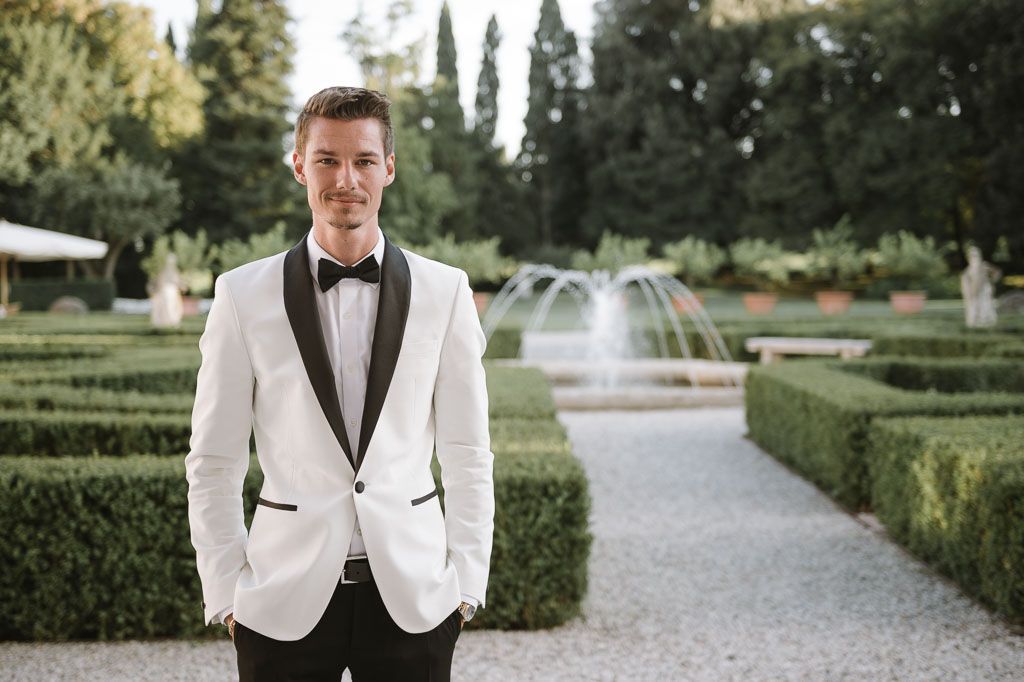 Groom in white tuxedo jacket poses in manicured garden with fountain backdrop.