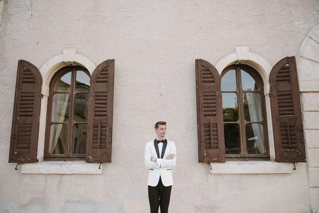 Classic groom portrait in white tuxedo standing between arched windows with brown shutters.