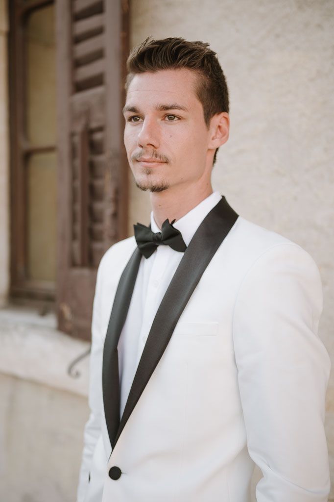 Sophisticated groom portrait in white dinner jacket and black bow tie against stone wall.